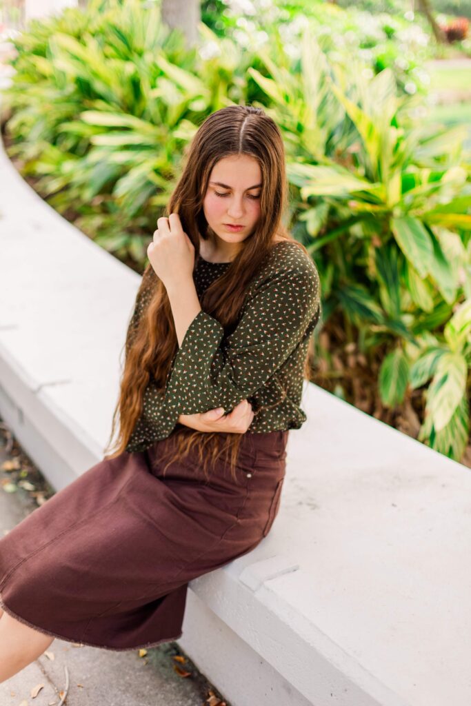 Girl sits on low wall and looks down during scenic portrait session in Orlando Florida.