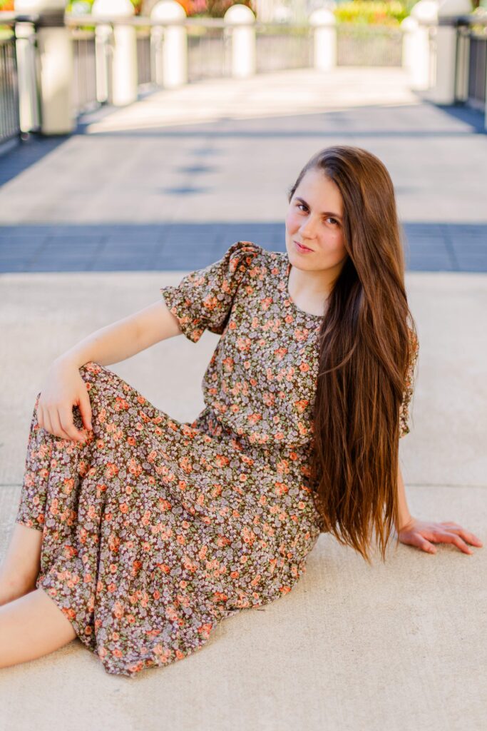 Girl sits on the ground and rests one hand on knee while looking at the camera during scenic portrait session in Orlando Florida.