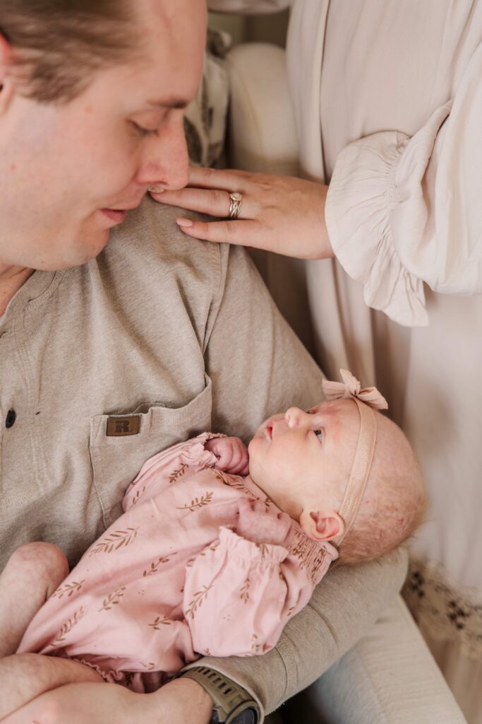 Mom rests her hand on Dad's shoulder while Dad holds baby during in home newborn session.