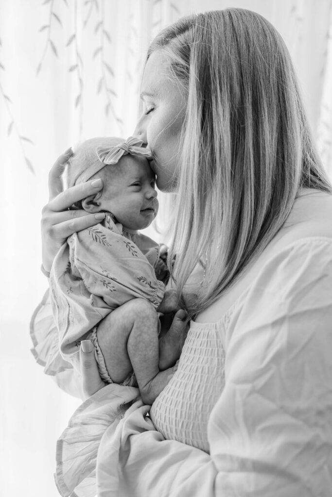 Mom holds newborn in front of the window and kisses her on the forehead.