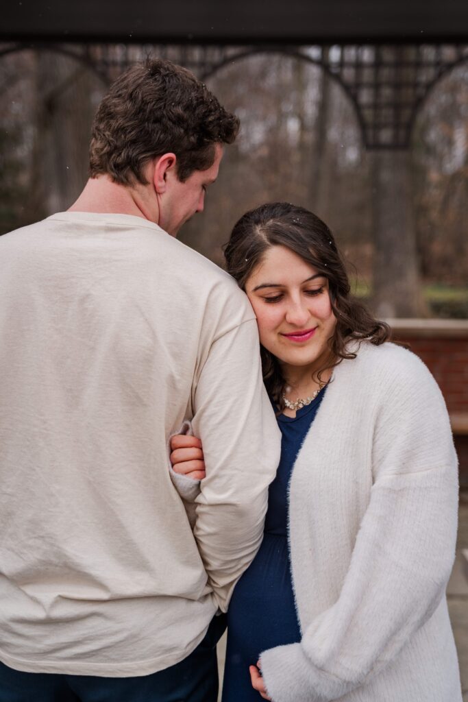 Wife looks down while leaning on husband's shoulder under an arbor during snowy maternity session.