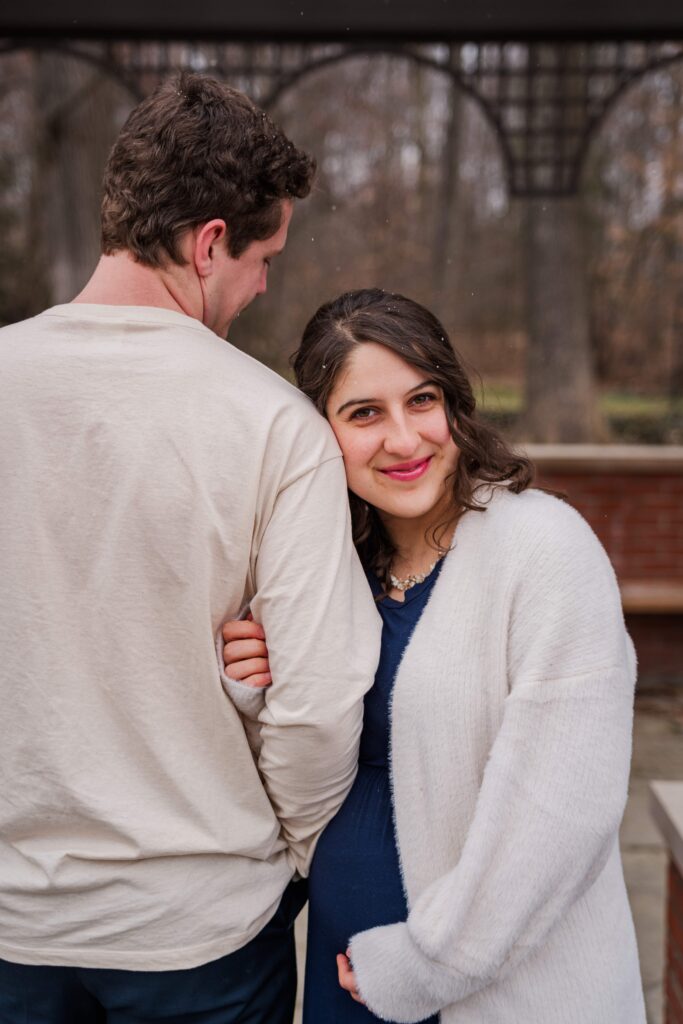 Wife smiles at the camera while leaning on husband's shoulder under an arbor.