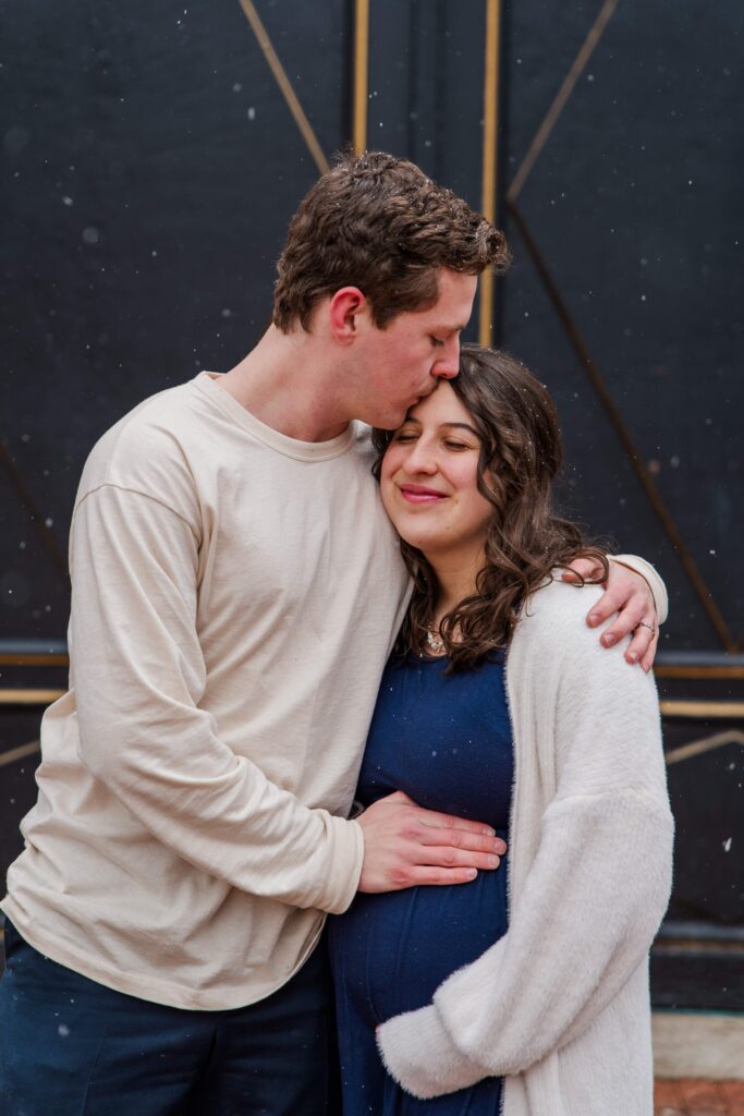 Husband and wife rest their hands on baby bump. Husband kisses wife.