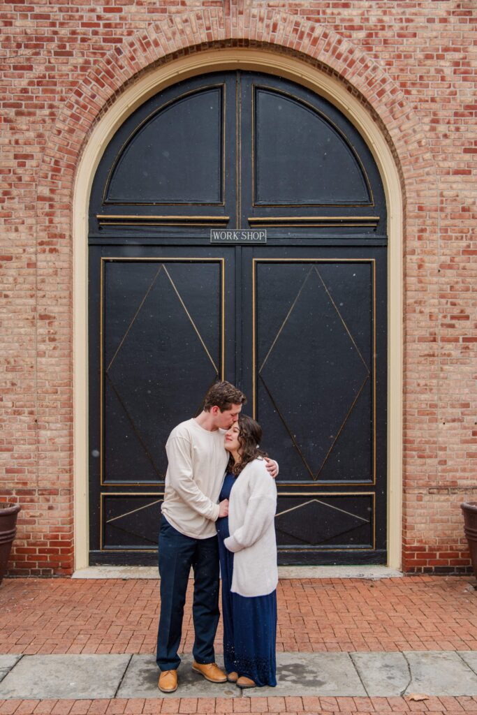 Husband and wife rest their hands on baby bump in front of a big door.