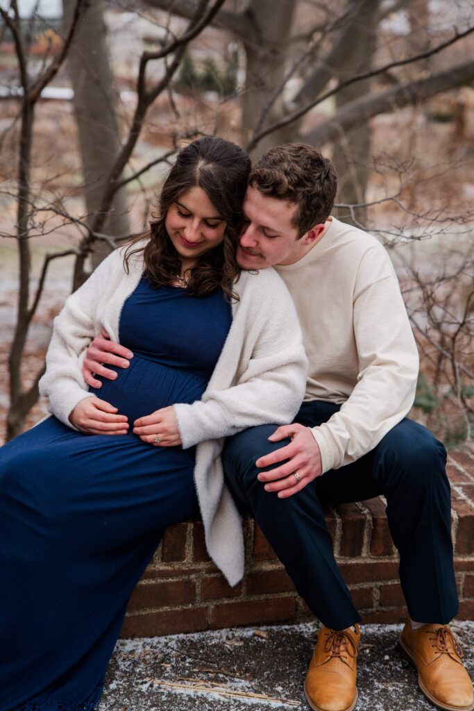 Couple smiling down at their baby bump while sitting on a low wall.