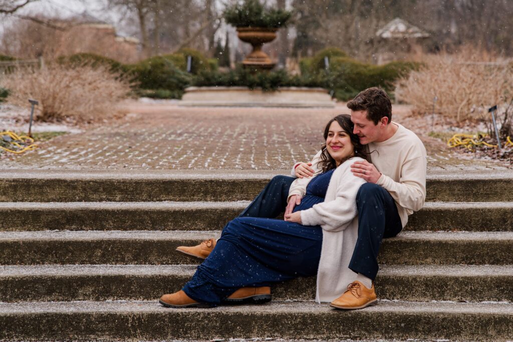 Couple sits on the stairs and smile while resting their hands on the baby bump at snowy maternity session