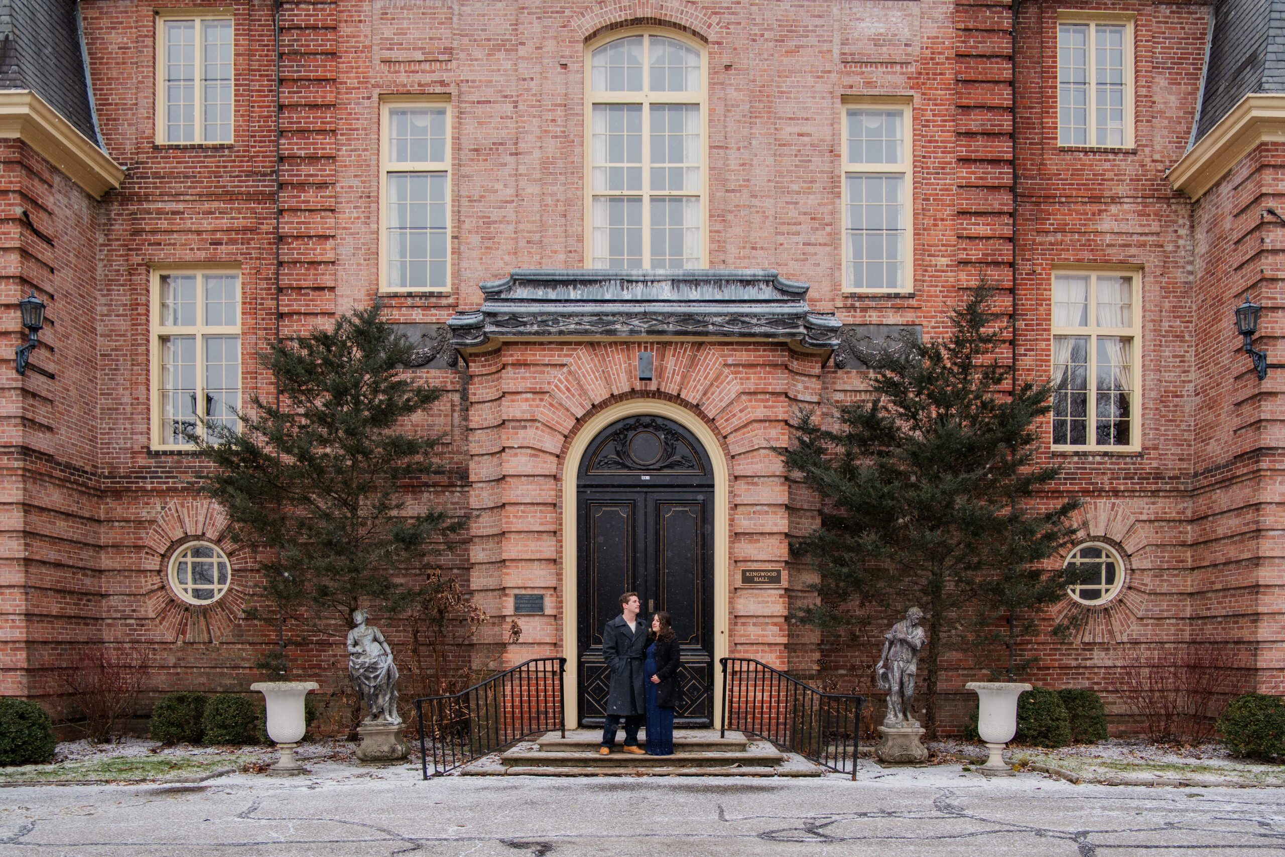 Couple stands in front of Kingwood Hall. Wife rests her hand on her baby bump.