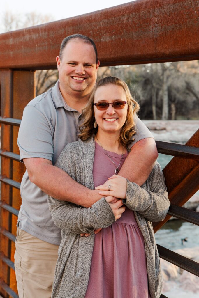 Dad and mom stand by bridge railings and smile at the camera.